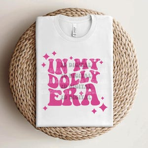 May include: A white t-shirt with the text "IN MY DOLLY ERA" in a retro pink font, accented with pink starburst graphics. The shirt is folded and placed on a woven, circular mat.