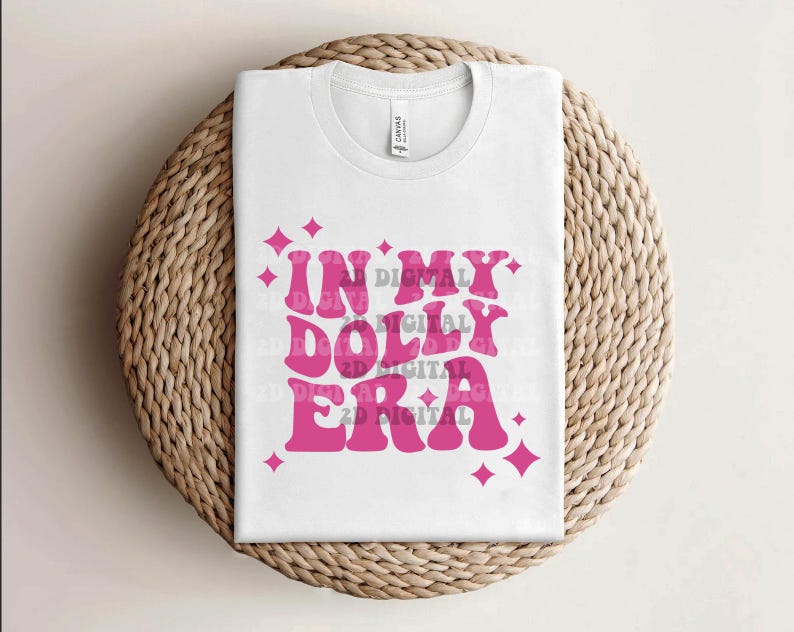 May include: A white t-shirt with the text "IN MY DOLLY ERA" in a retro pink font, accented with pink starburst graphics. The shirt is folded and placed on a woven, circular mat.