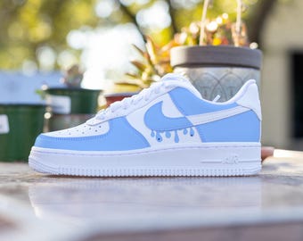 May include: White and light blue sneakers with a blue swoosh logo and blue paint drips. The sneakers have a white midsole and a white outsole.