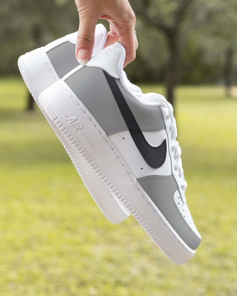 May include: Gray and white low-top sneakers with a black Nike swoosh logo. The sneakers are held in a hand and the background is blurred.