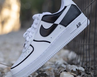 May include: White leather low-top sneakers with a black leather swoosh and black leather panel on the back. The sneakers have a black outline and the word "AIR" is printed on the side.