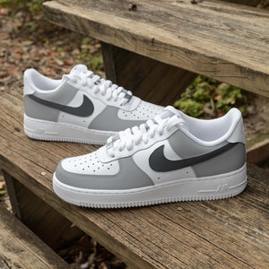 May include: A pair of white and gray Nike Air Force 1 sneakers with a black swoosh logo. The sneakers are on a wooden surface.