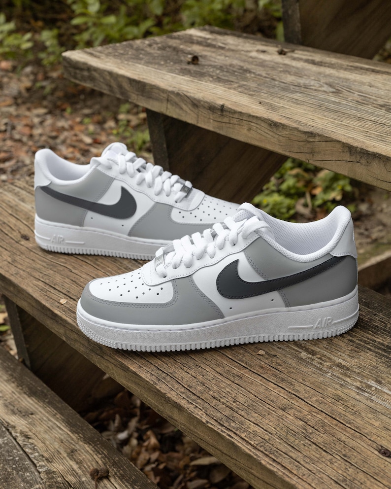 May include: A pair of white and gray Nike Air Force 1 sneakers with a black swoosh logo. The sneakers are on a wooden surface.