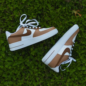 May include: A pair of white trainers with brown accents. The trainers have a brown swoosh logo and a brown stripe across the toe. The trainers are on a green grassy surface.
