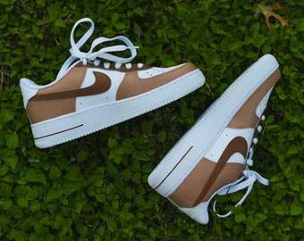 May include: A pair of white sneakers with brown accents. The sneakers have a brown swoosh logo and a brown stripe across the toe. The sneakers are on a green grassy surface.