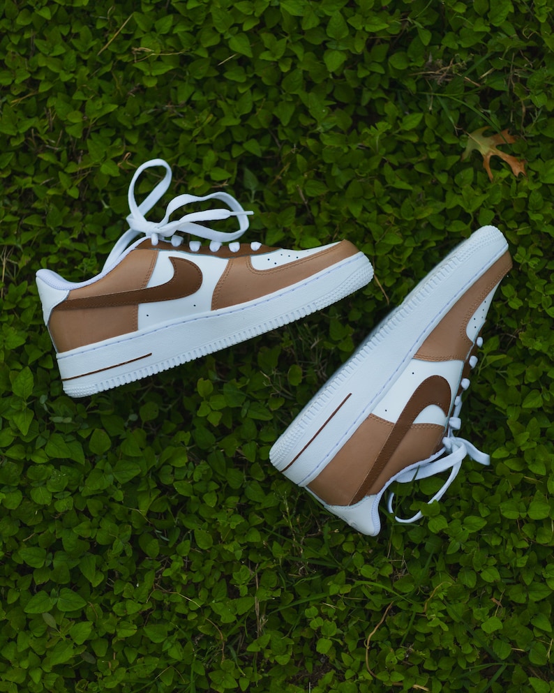 May include: A pair of white trainers with brown accents. The trainers have a brown swoosh logo and a brown stripe across the toe. The trainers are on a green grassy surface.