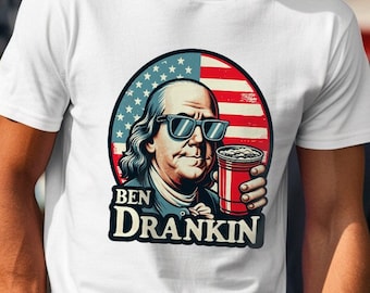Puede incluir: Camiseta blanca con un gráfico de Benjamin Franklin con gafas de sol y sosteniendo una taza roja. El gráfico está rodeado por un diseño de bandera estadounidense. El texto "BEN DRANKIN" está debajo del gráfico.