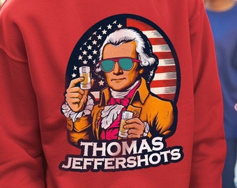 Puede incluir: Sudadera roja con un gráfico de Thomas Jefferson con gafas de sol y sosteniendo dos vasos de chupito. El gráfico está rodeado por un diseño de bandera estadounidense. El texto "Thomas Jeffershots" está impreso debajo del gráfico.
