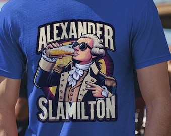 Puede incluir: Una camiseta azul con una imagen de dibujos animados de Alexander Hamilton con gafas de sol y bebiendo de un vaso. El texto "Alexander Slamilton" está impreso debajo de la imagen.