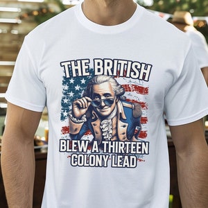 May include: A white t-shirt with a graphic of a man wearing sunglasses and a tri-cornered hat in front of an American flag. The text on the shirt reads "The British Blew a Thirteen Colony Lead."