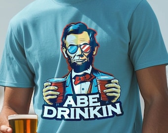 May include: A light blue t-shirt with a cartoon illustration of Abraham Lincoln wearing sunglasses and holding two red cups. The text "Abe Drinkin'" is printed below the illustration.
