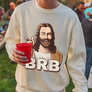 Könnte beinhalten: Ein weißer Pullover mit einem Cartoon-Bild von Jesus, der eine rote Tasse hält, und dem Text "BRB".