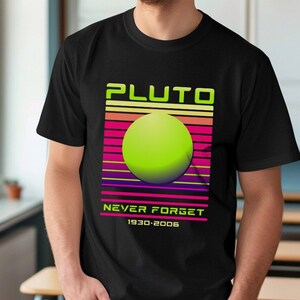 May include: Black t-shirt with a retro design featuring the word "PLUTO" in neon green, a green sphere, and the text "NEVER FORGET 1930-2006". The shirt is worn by a person wearing glasses and blue jeans.
