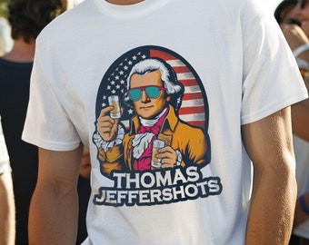 Puede incluir: Camiseta blanca con un gráfico de Thomas Jefferson con gafas de sol y bebidas, con el texto "THOMAS JEFFERSHOTS". La persona lleva una gorra de béisbol negra con una bandera estadounidense.