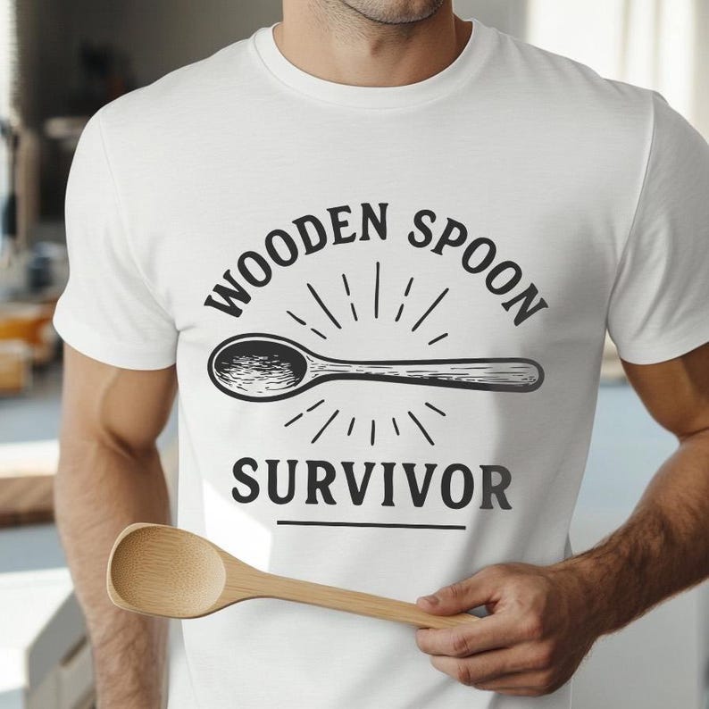 Op de afbeelding: Wit T-shirt met een zwarte afbeelding van een houten lepel en de tekst "WOODEN SPOON SURVIVOR". Een houten lepel wordt op de voorgrond gehouden.