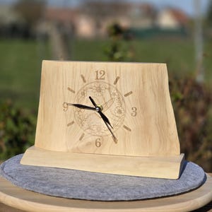 Könnte beinhalten: Helle Holz-Uhr mit floralem Design um die Ziffern. Die Uhr hat schwarze Zeiger und steht auf einem Holzsockel. Die Uhr wird auf einem grauen Filzpad präsentiert. Schlichtes und elegantes Design.