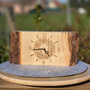 Könnte beinhalten: Eine Holzuhr mit natürlichem Rindenrand. Das Zifferblatt hat ein Blumendesign mit schwarzen Zeigern und Zahlen. Die Uhr ist aus hellem Holz gefertigt und steht auf einem grauen Filzpad.