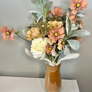 Könnte beinhalten: Ein Strauß künstlicher Blumen in einer durchsichtigen Glasvase, gefüllt mit braunem Sand. Der Strauß enthält weiße, cremefarbene und pfirsichfarbene Blumen mit grünem Laub.