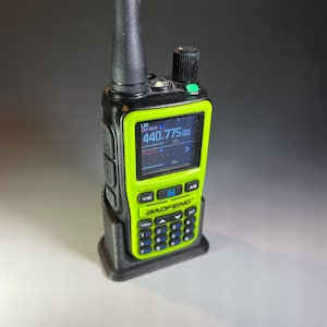 May include: A handheld Baofeng radio in a black charging cradle. The radio has a bright green frame with a black antenna and buttons. The screen displays numbers and letters. The radio is designed for communication.