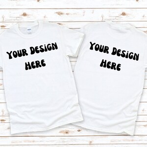 Peut inclure: Deux t-shirts blancs avec le texte "YOUR DESIGN HERE" en gras noir. Les chemises sont présentées sur une surface à grain de bois clair, montrant le devant et le dos des chemises. Les chemises sont vierges, prêtes à être personnalisées.