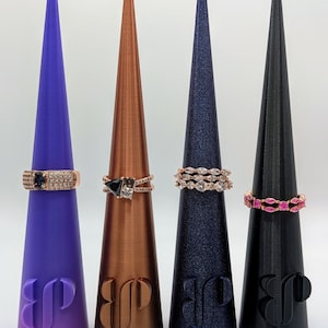 May include: Four ring holders in purple, bronze, navy, and black, each with a different ring. The rings feature various gemstones and designs. The holders have a logo at the base.