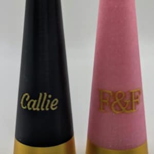 May include: Two decorative, cone-shaped objects. One is black with the name "Callie" in gold script. The other is pink with gold glitter and the letters "F & F". Both have gold-colored bases.