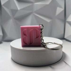 May include: A pink, cube-shaped keychain with the initials "BP" carved into one side. The keychain is attached to a silver-colored chain and ring. The background is a white, geometric pattern.