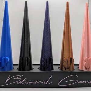 May include: Five cone-shaped objects in blue, black, purple, bronze, and pink, each with a logo, displayed in a black tray. The tray has the text "Botanical Gems" in a cursive font.