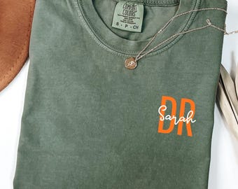 May include: Moss green t-shirt with the text "DR Sarah" in orange. The shirt is folded and has a Comfort Colors tag. A gold necklace with a pendant is draped across the shirt. A brown hat is visible in the upper left corner.