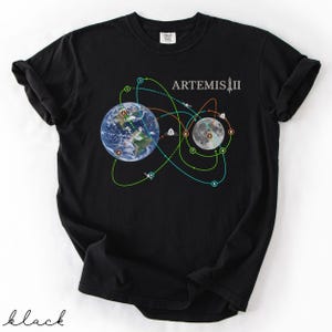 May include: Black t-shirt featuring a graphic of Earth and the Moon with a flight path design, labeled "ARTEMIS II." The design includes colorful lines and numbered circles. The word "black" is written in cursive at the bottom.