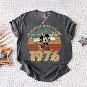 May include: Dark grey t-shirt with a vintage Mickey Mouse design celebrating a 50th anniversary. The design includes the text "50 Years of Being Awesome" and "1976 Limited Edition". The shirt has rolled sleeves and a vintage look.