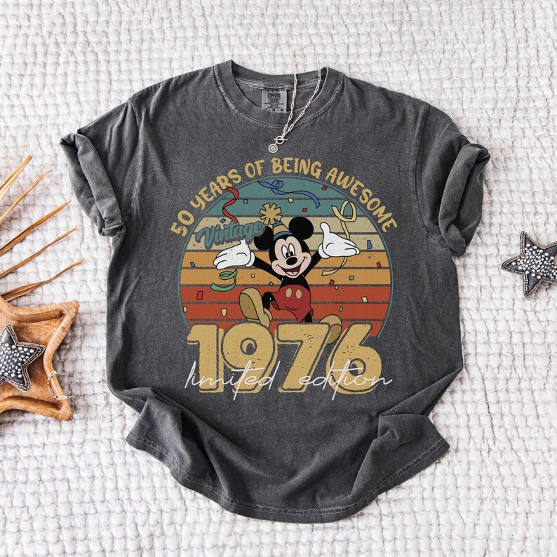 May include: Dark grey t-shirt with a vintage Mickey Mouse design celebrating a 50th anniversary. The design includes the text "50 Years of Being Awesome" and "1976 Limited Edition". The shirt has rolled sleeves and a vintage look.