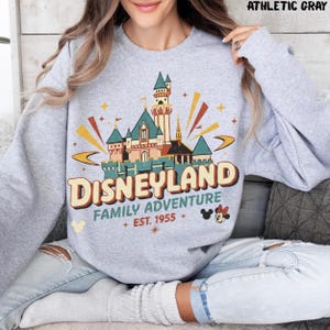 Puede incluir: Sudadera gris atl&eacute;tica con un gr&aacute;fico colorido del castillo de Disneyland. El gr&aacute;fico incluye el texto "Disneyland Family Adventure + Est. 1955" con iconos de Mickey y Minnie Mouse. La sudadera es de cuello redondo.