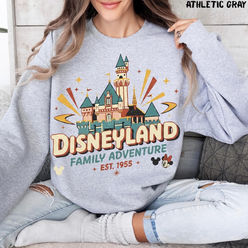 Puede incluir: Sudadera gris atl&eacute;tica con un gr&aacute;fico colorido del castillo de Disneyland. El gr&aacute;fico incluye el texto "Disneyland Family Adventure + Est. 1955" con iconos de Mickey y Minnie Mouse. La sudadera es de cuello redondo.