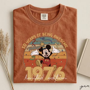 May include: A terracotta-coloured t-shirt with a Mickey Mouse graphic celebrating. The design includes the text "50 YEARS OF BEING AWESOME" and "1976 limited edition" in a vintage style. The shirt has a crew neck and a Comfort Colors tag.