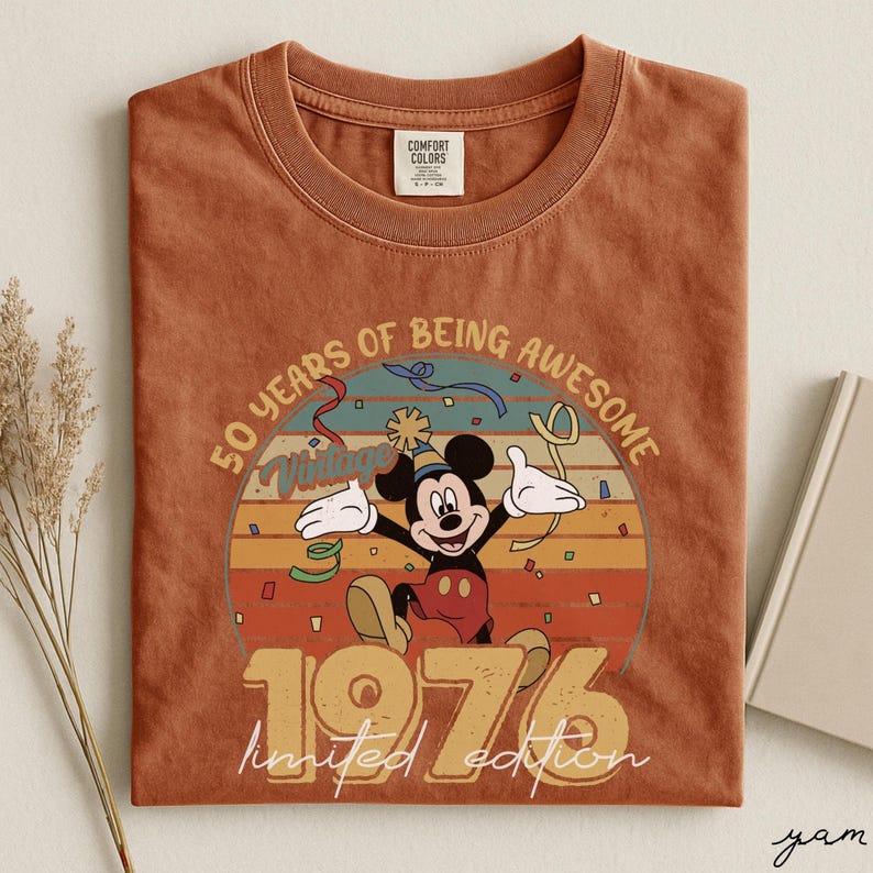 May include: A terracotta-coloured t-shirt with a Mickey Mouse graphic celebrating. The design includes the text "50 YEARS OF BEING AWESOME" and "1976 limited edition" in a vintage style. The shirt has a crew neck and a Comfort Colors tag.