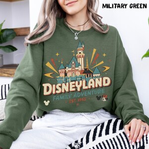 Puede incluir: Sudadera verde militar con un gr&aacute;fico del castillo de Disneyland. El gr&aacute;fico incluye la palabra "Disneyland", "Family Adventure" y "Est. 1955". La sudadera tiene cuello redondo y mangas largas.