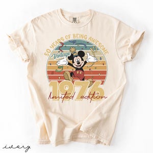 May include: Ivory t-shirt with a vintage Mickey Mouse design celebrating 50 years. The graphic includes the text "50 Years of Being Awesome," "Vintage," and "1976 Limited Edition" in a retro style, with a circular background.