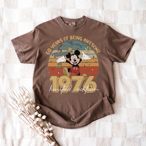 May include: Brown t-shirt with a Mickey Mouse graphic celebrating a milestone. The design includes the text "50 years of being awesome" and "1976 limited edition". The shirt has a vintage, retro style.