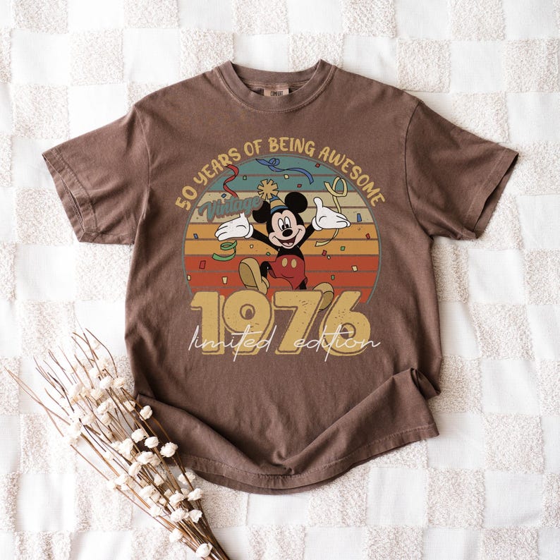 May include: Brown t-shirt with a Mickey Mouse graphic celebrating a milestone. The design includes the text "50 years of being awesome" and "1976 limited edition". The shirt has a vintage, retro style.