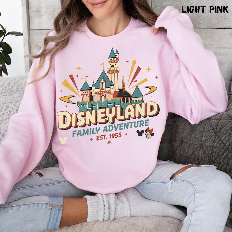 Puede incluir: Sudadera rosa claro con un dise&ntilde;o de Disneyland. El dise&ntilde;o presenta un castillo, las palabras "Disneyland Family Adventure" y el a&ntilde;o "Est. 1955". Tambi&eacute;n se incluyen iconos de Mickey y Minnie Mouse.