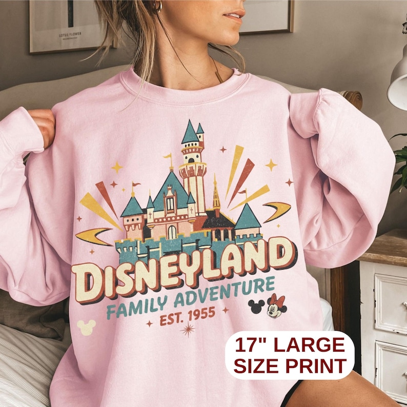 Puede incluir: Una sudadera rosa claro con un gr&aacute;fico de Disneyland. El gr&aacute;fico presenta un castillo, la palabra "Disneyland" y el texto "Family Adventure + Est. 1955 +" con una impresi&oacute;n grande de 43 cm.