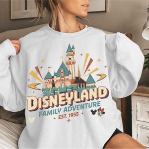 Puede incluir: Sudadera blanca con un gr&aacute;fico de Disneyland. El dise&ntilde;o incluye un castillo, la palabra "Disneyland" y la frase "Family Adventure Est. 1955". Mickey Mouse y estrellas tambi&eacute;n est&aacute;n incluidas en el dise&ntilde;o.