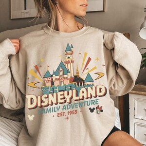 Puede incluir: Sudadera beige de cuello redondo con un gr&aacute;fico colorido de Disneyland. El dise&ntilde;o incluye un castillo, la palabra "Disneyland", "Family Adventure" y "Est. 1955". Mickey Mouse y Minnie Mouse tambi&eacute;n est&aacute;n presentes.
