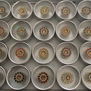 May include: A set of 18 small round containers with lids, each containing a decorative jeweled flower embellishment. The embellishments are in various colours, including red, green, blue, gold, and white.