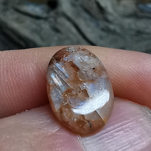 May include: An oval-shaped gemstone with a polished surface. The stone has a translucent quality, displaying a blend of brown, white, and clear inclusions. The gem is held in a hand, highlighting its size and intricate details.