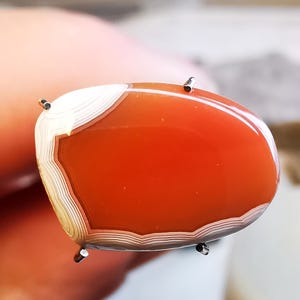 以下が含まれることがあります： 白い縞模様のある赤い瑪瑙のカボションのクローズアップ。石は4本の金属製の爪で固定されています。