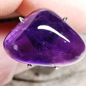 May include: A polished, deep purple gemstone with a rounded, triangular shape. The stone is mounted with small silver prongs. The surface reflects light, creating a bright white curved highlight. The gemstone is likely an amethyst.