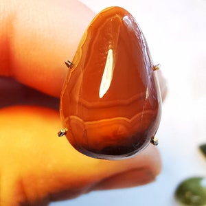 以下が含まれることがあります： 茶色とオレンジの縞模様が入った涙型の瑪瑙のカボション。石は銀製の爪付きベゼルにセットされています。