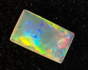 May include: A rectangular, translucent opal gemstone with a white base and iridescent flashes of blue, green, yellow, and red.